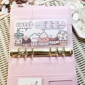 Op de afbeelding: Een roze kunstleren binder met goudkleurige hardware, open om een ​​transparante zak met een "Sweet Escape"-ontwerp te laten zien. Het ontwerp bevat cartoon illustraties van desserts en de tekst "Sweet Escape".