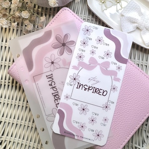 Può includere: Un'agenda rosa con un design floreale e la scritta "stay inspired". L'agenda include una sovrapposizione trasparente con un motivo floreale e una copertina rosa simile alla pelle. Elementi decorativi: un vassoio bianco con clip a forma di fiocco dorato.