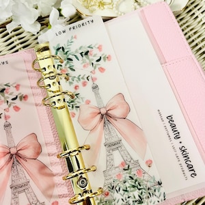 May include: A pink planner with gold rings, decorated with Eiffel Tower illustrations, pink bows, and floral details. The planner includes the text "LOW PRIORITY" and "beauty + skincare" with related product categories.