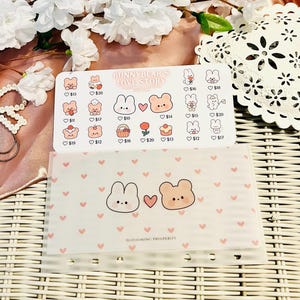 Può includere: Una busta per budget in plastica trasparente con un motivo a cuori rosa e illustrazioni di un coniglietto e un orsetto. È visibile anche un foglio di adesivi con il testo "BUNNYBEAR'S LOVE STORY".