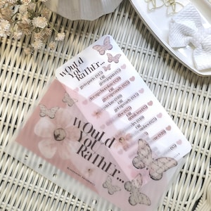May include: Two Would You Rather game cards with butterfly and floral designs. The cards have various choices like "go to Paris" or "give up candy." A white bow and gold butterfly hair clips are on a white tray.