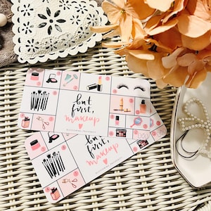 May include: Two pink and white cards with a makeup theme. The cards have a grid of makeup items and the text "but first, makeup" with a heart. The cards are on a woven wicker surface.