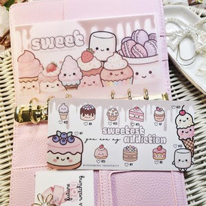 Op de afbeelding: Een roze planner met gouden ringen, met decoratieve inzetstukken met dessert-thema illustraties. De inzetstukken bevatten afbeeldingen van cupcakes, macarons en taarten, met de woorden "sweet" en "sweetest addiction".