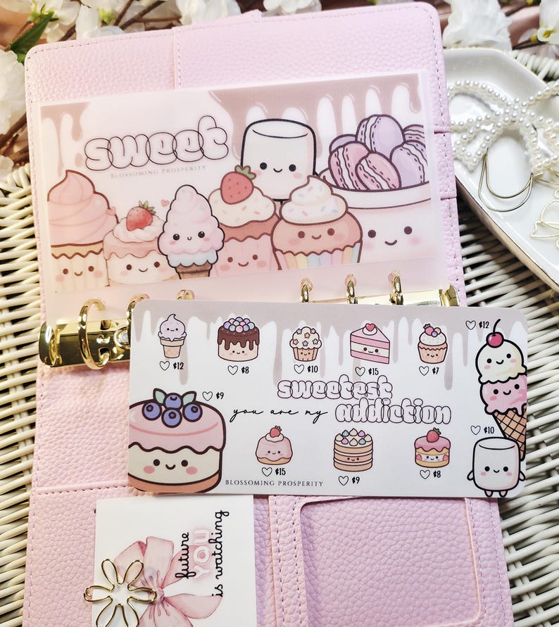 Op de afbeelding: Een roze planner met gouden ringen, met decoratieve inzetstukken met dessert-thema illustraties. De inzetstukken bevatten afbeeldingen van cupcakes, macarons en taarten, met de woorden "sweet" en "sweetest addiction".