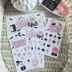 May include: A collection of white sticker sheets featuring pink and black illustrations of ghosts, hearts, bats, bows, and magical items. Text includes Boo-tiful, Haunted Hearts, and Spooky Spells. A small black card with the text Let every choice reflect your values.