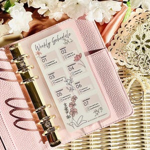 Peut inclure: Un agenda rose avec un insert de planning hebdomadaire. L'insert présente un motif floral et un emploi du temps pour chaque jour de la semaine, avec des activités comme le dîner, le travail et les rendez-vous au spa. L'agenda est ouvert, révélant des anneaux dorés.