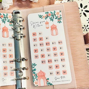 May include: A pink faux leather binder with a savings tracker insert. The insert features a floral design with pink lanterns and the text "Shine from Within." It includes spaces to track savings goals, with amounts from $1 to $10.
