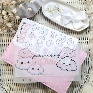 Peut inclure: Un agenda rose avec un recouvrement transparent orné d'illustrations de nuages et du texte "just chasing CLOUDS". Le recouvrement comprend également des autocollants de nuages avec des symboles monétaires. Un plateau blanc contient des clips en forme de nœuds dorés et un nœud blanc.