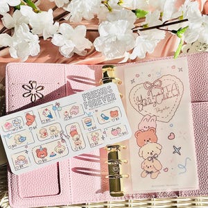 May include: A pink planner with gold-coloured hardware, open to show a sticker sheet and a decorative page. The sticker sheet has cartoon bears, ice cream, and other images. The decorative page has a heart-shaped design with cartoon animals.
