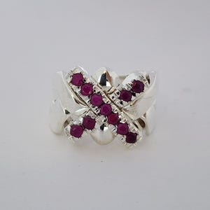 Puede incluir: Un anillo de plata con un diseño ancho de varias bandas y una pieza central prominente en forma de X. El elemento cruzado está engastado con pequeñas piedras preciosas redondas de color rojo rosado intenso, alineadas a lo largo de las dos barras.