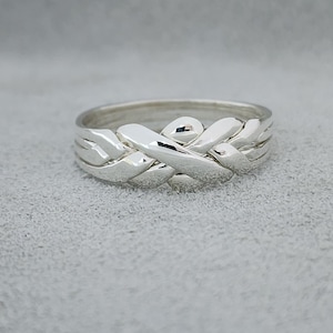 May include: A polished silver puzzle ring featuring four interlocking bands that weave together to form a single cohesive unit. The metal surface is smooth and reflective, showcasing a complex, braided design with rounded edges and a continuous loop.