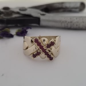 May include: A gold puzzle ring featuring four interlocking bands, with two of the bands forming an X-shaped centerpiece set with six small, round, deep red rubies. The ring has a polished metallic finish and a complex, woven design structure.