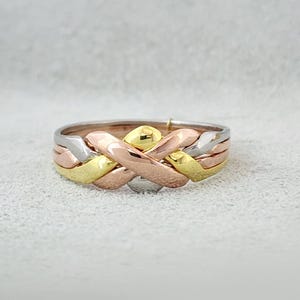 May include: A tri-colour puzzle ring featuring four interlocking bands in yellow, rose, and white gold tones. The bands are woven together in a complex, overlapping pattern that creates a continuous, unified circular shape for the finger.