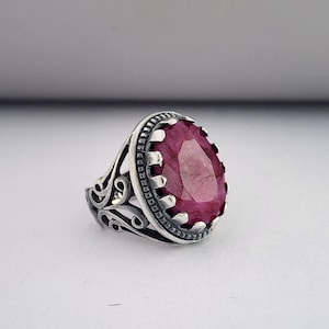 Peut inclure: Une bague en argent avec un design orné présente une grande pierre précieuse ovale de couleur rose foncé.