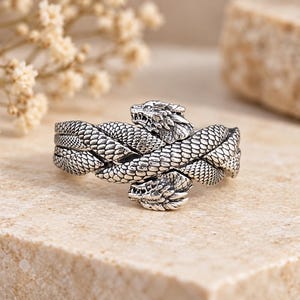 Puede incluir: Un anillo de tono plateado que presenta dos cabezas de dragón entrelazadas con escamas detalladas. El metal tiene un acabado oxidado que resalta la textura de los cuerpos de los dragones y los rasgos afilados de las cabezas, creando un diseño continuo.