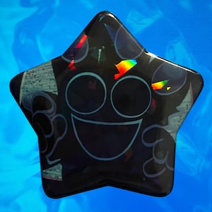 May include: A black, star-shaped object with a cartoon face design. The face features large, round eyes, a wide smile, and a light blue outline. The object has a glossy finish with iridescent rainbow accents.