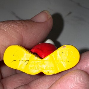 K&ouml;nnte beinhalten: Kleine, alte Figur einer Figur mit rotem Kopf, wei&szlig;em Hut und gelben F&uuml;&szlig;en. Die Figur wird in einer Hand gehalten, wobei die Unterseite sichtbar ist und den Text "&copy;Disney MADE IN CHINA" anzeigt.
