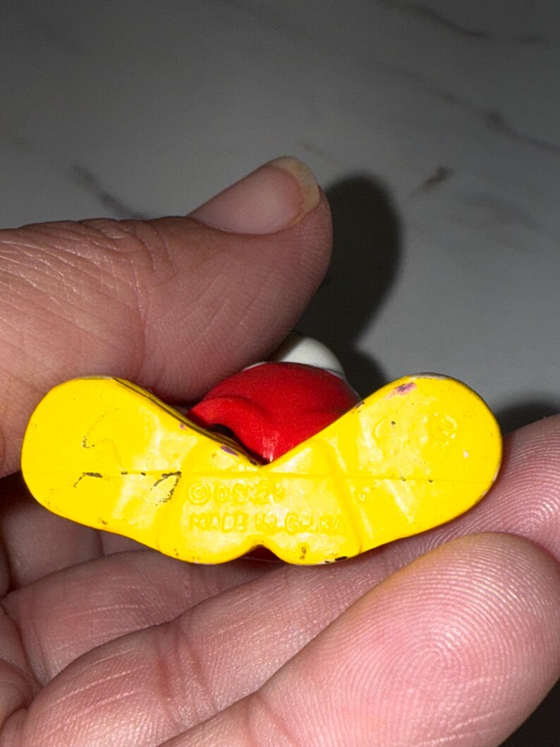 K&ouml;nnte beinhalten: Kleine, alte Figur einer Figur mit rotem Kopf, wei&szlig;em Hut und gelben F&uuml;&szlig;en. Die Figur wird in einer Hand gehalten, wobei die Unterseite sichtbar ist und den Text "&copy;Disney MADE IN CHINA" anzeigt.