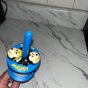 May include: A blue and yellow spinning top with a Mickey Mouse design. The top has two Mickey Mouse faces on the side and the word "Mickey" written on the bottom.