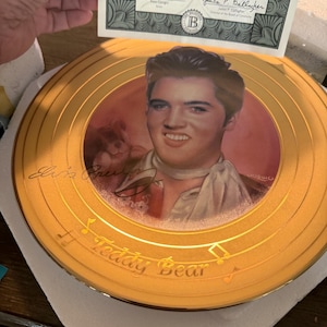 May include: A decorative gold-toned plate featuring a portrait of Elvis Presley. The plate has a gold rim and the words "Teddy Bear" are inscribed. The plate is accompanied by a certificate from The Bradford Exchange.