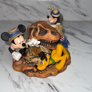 May include: A Disney figurine featuring Mickey Mouse, Goofy, and Pluto in a dinosaur-themed setting. Mickey and Goofy wear safari hats, while Pluto lies on the ground. The figurine includes a dinosaur skull and a sign that reads "Dino-Rama".