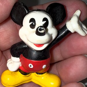 K&ouml;nnte beinhalten: Kleine Mickey Mouse-Figur. Die Figur hat ein wei&szlig;es Gesicht, schwarze Ohren und einen roten Mund. Sie tr&auml;gt rote Shorts, gelbe Schuhe und hat schwarze Arme. Ein Arm ist zum Winken erhoben. Ein klassisches Disney-Sammlerst&uuml;ck.
