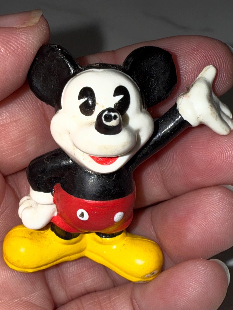 K&ouml;nnte beinhalten: Kleine Mickey Mouse-Figur. Die Figur hat ein wei&szlig;es Gesicht, schwarze Ohren und einen roten Mund. Sie tr&auml;gt rote Shorts, gelbe Schuhe und hat schwarze Arme. Ein Arm ist zum Winken erhoben. Ein klassisches Disney-Sammlerst&uuml;ck.