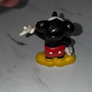 K&ouml;nnte beinhalten: Eine kleine Mickey Mouse-Figur. Die Figur ist schwarz mit wei&szlig;en Handschuhen, einem wei&szlig;en Gesicht und roten Shorts. Die Schuhe sind gelb. Die Figur posiert mit einem erhobenen Arm.