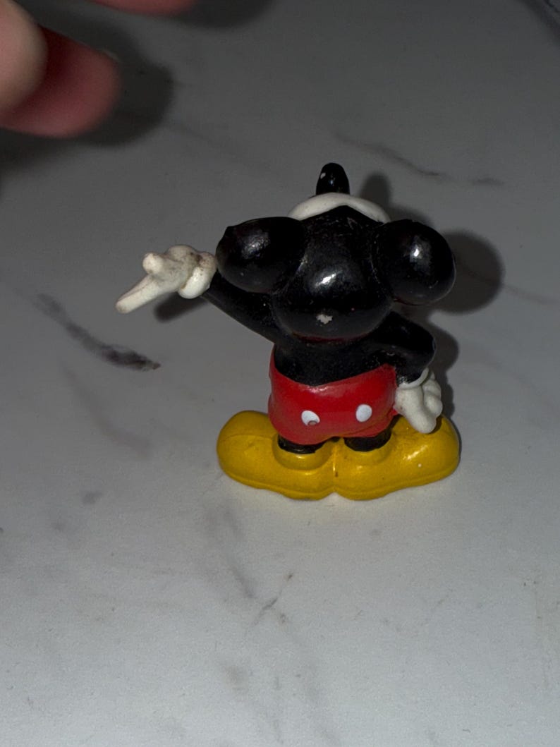 K&ouml;nnte beinhalten: Eine kleine Mickey Mouse-Figur. Die Figur ist schwarz mit wei&szlig;en Handschuhen, einem wei&szlig;en Gesicht und roten Shorts. Die Schuhe sind gelb. Die Figur posiert mit einem erhobenen Arm.