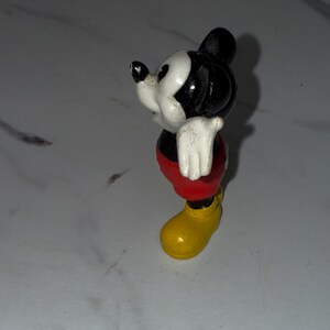 K&ouml;nnte beinhalten: Eine kleine Mickey Mouse-Figur. Die Figur hat einen schwarzen Kopf und Ohren, ein wei&szlig;es Gesicht, wei&szlig;e Handschuhe, rote Shorts und gelbe Schuhe. Die Figur steht auf einer wei&szlig;en Oberfl&auml;che.