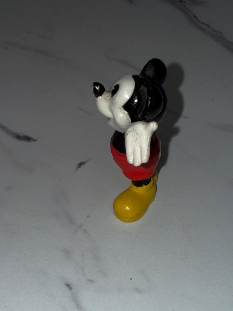 K&ouml;nnte beinhalten: Eine kleine Mickey Mouse-Figur. Die Figur hat einen schwarzen Kopf und Ohren, ein wei&szlig;es Gesicht, wei&szlig;e Handschuhe, rote Shorts und gelbe Schuhe. Die Figur steht auf einer wei&szlig;en Oberfl&auml;che.