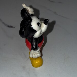 K&ouml;nnte beinhalten: Eine Vintage-Mickey-Mouse-Figur. Die Figur ist haupts&auml;chlich schwarz und wei&szlig;, mit roten Shorts und gelben Schuhen. Die Figur posiert mit einem erhobenen Arm und dem anderen an der Seite.