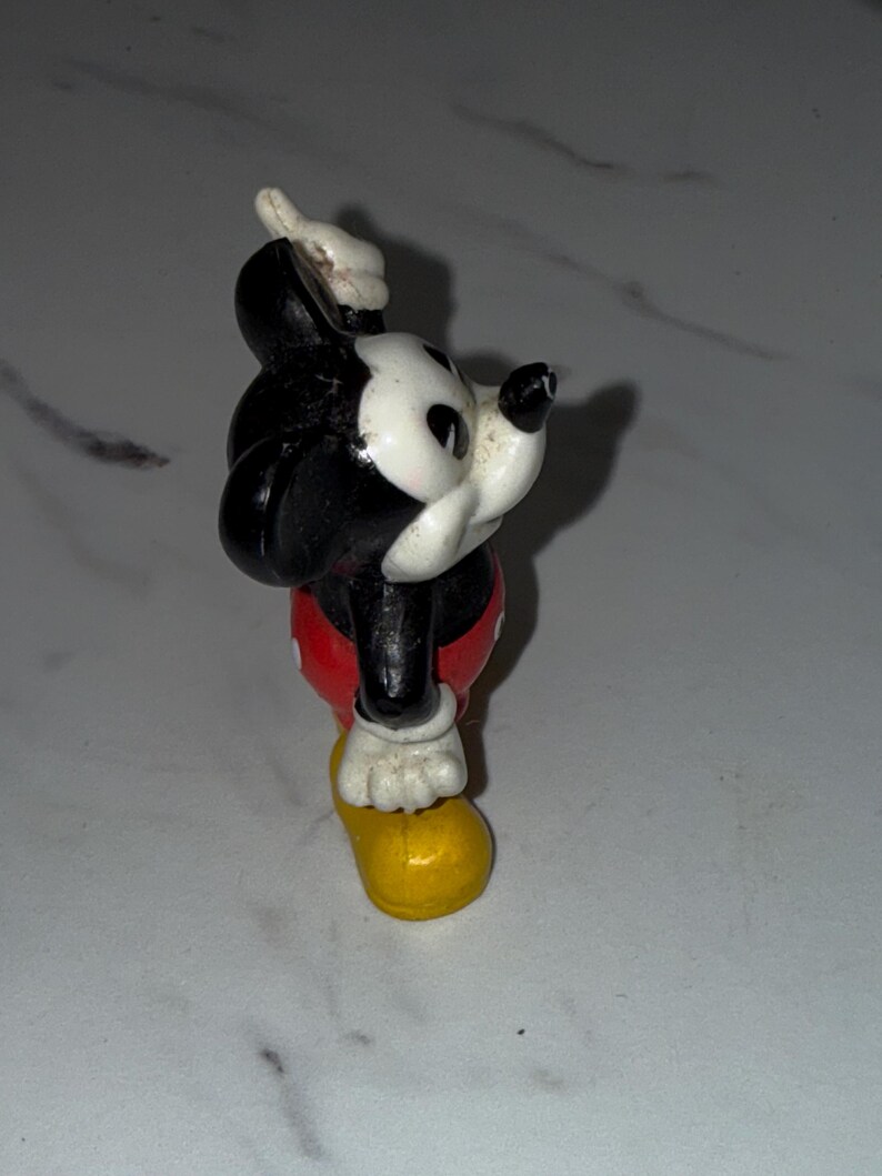 K&ouml;nnte beinhalten: Eine Vintage-Mickey-Mouse-Figur. Die Figur ist haupts&auml;chlich schwarz und wei&szlig;, mit roten Shorts und gelben Schuhen. Die Figur posiert mit einem erhobenen Arm und dem anderen an der Seite.