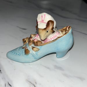 May include: A blue ceramic shoe with a mother mouse and two baby mice inside. The mother mouse is wearing a pink and white hat and dress.