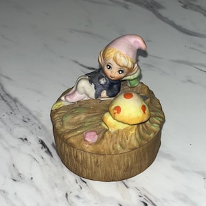 May include: A ceramic trinket box featuring a whimsical gnome figure in a pink hat and a yellow mushroom. The box is round with a textured, brown exterior, and the gnome is lying down.