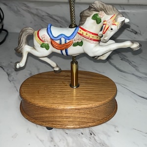 May include: A decorative carousel horse figurine. The white horse has a gray mane and tail, adorned with pink flowers and a red and gold saddle. It is mounted on a wooden base with a brass pole.