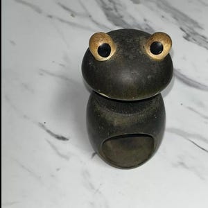 May include: A dark green frog-shaped ring holder with large, gold-colored eyes. The ring holder is made of wood and has a smooth, rounded shape. The frog's eyes are detailed with black pupils. The ring holder is designed to hold rings.