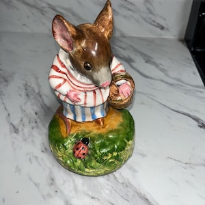 May include: A ceramic figurine of a brown mouse wearing a red and blue striped shirt, holding a basket and standing on a green base with a ladybug.