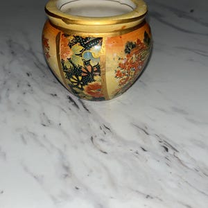 May include: A small, round ceramic pot with a wide opening. The pot features a gold rim and is decorated with floral and landscape designs in shades of orange, yellow, and black. The interior is white.