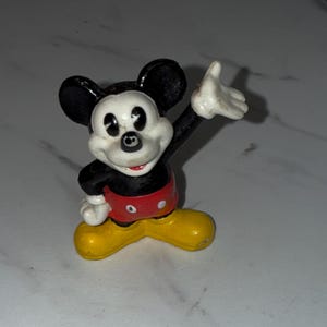 K&ouml;nnte beinhalten: Eine kleine, alte Mickey Mouse-Figur. Die Figur befindet sich in einer klassischen Pose, mit einem erhobenen Arm. Mickey tr&auml;gt eine schwarze Hose, ein rotes Oberteil, gelbe Schuhe und wei&szlig;e Handschuhe. Die Figur hat schwarze Ohren und ein wei&szlig;es Gesicht.