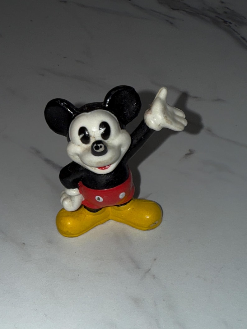 K&ouml;nnte beinhalten: Eine kleine, alte Mickey Mouse-Figur. Die Figur befindet sich in einer klassischen Pose, mit einem erhobenen Arm. Mickey tr&auml;gt eine schwarze Hose, ein rotes Oberteil, gelbe Schuhe und wei&szlig;e Handschuhe. Die Figur hat schwarze Ohren und ein wei&szlig;es Gesicht.
