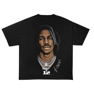 May include: Black t-shirt featuring a portrait of a person with dreadlocks and multiple silver chain necklaces. The number "12" is below the portrait, along with a signature. The shirt is a solid black color.