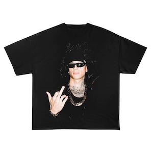 Puede incluir: Camiseta negra con una persona que lleva gafas de sol, una bandana negra y una gruesa cadena. La persona hace un gesto con la mano. La imagen está sobre un fondo oscuro.