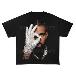 May include: Black t-shirt featuring a portrait of a man with a diamond-studded glove making an 'okay' gesture. The image is a close-up, highlighting the man's face, jewelry, and the intricate details of the glove. The shirt is a classic crew neck.