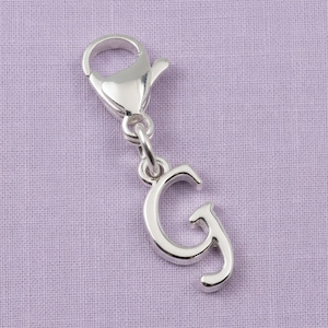 May include: A silver-toned metal charm in the shape of the uppercase letter G, featuring a smooth, reflective surface and a lobster claw clasp attached to the top, allowing it to be easily clipped onto various items like bracelets or necklaces.