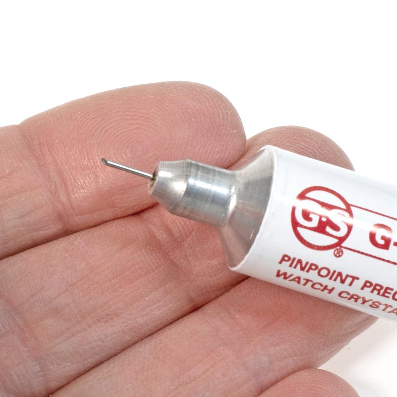Puede incluir: Un aplicador de cristal de reloj plateado y blanco con una punta de aguja. El aplicador tiene una etiqueta roja con el texto "GS PINPOINT PRECISION WATCH CRYSTALS".