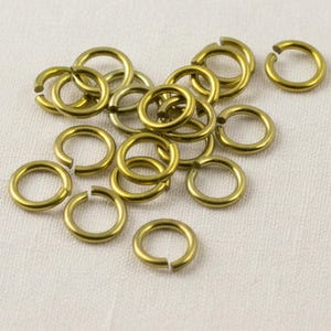 May include: A collection of small, circular metal jump rings with a matte gold finish. Each ring features a single open cut, allowing for easy connection and assembly in jewelry making projects. The rings are scattered in a loose pile.