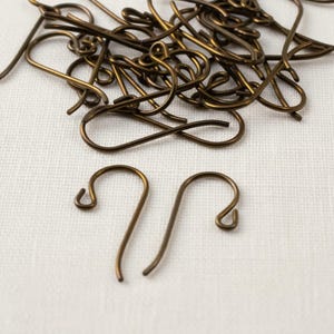 May include: A collection of bronze-colored earring hooks, some in a pile and two displayed separately. The hooks have a simple, curved design with a loop at the top and a curved bottom. The background is a white, textured fabric.