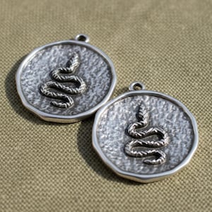 May include: Two silver-toned pendants with a textured surface and a raised snake design. Each pendant is circular with an irregular edge and a small loop at the top for hanging. The snake is coiled in a stylized manner.