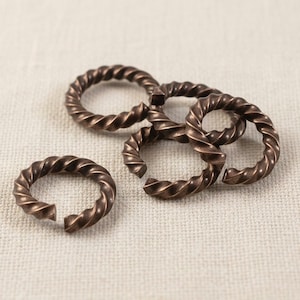May include: Close-up of several bronze-colored, twisted metal rings. The rings are open, with a gap on one side. The texture of the metal is visible, with a spiral pattern. The rings are scattered on a neutral-colored fabric.